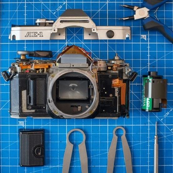 Appareil photo argentique Canon AE-1 démonté, outils et pellicule disposés sur une grille bleue.