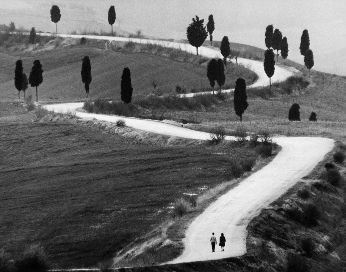 Route sinueuse en Toscane entourée de cyprès, avec deux personnes marchant côte à côte, photographie en noir et blanc de 1965.
