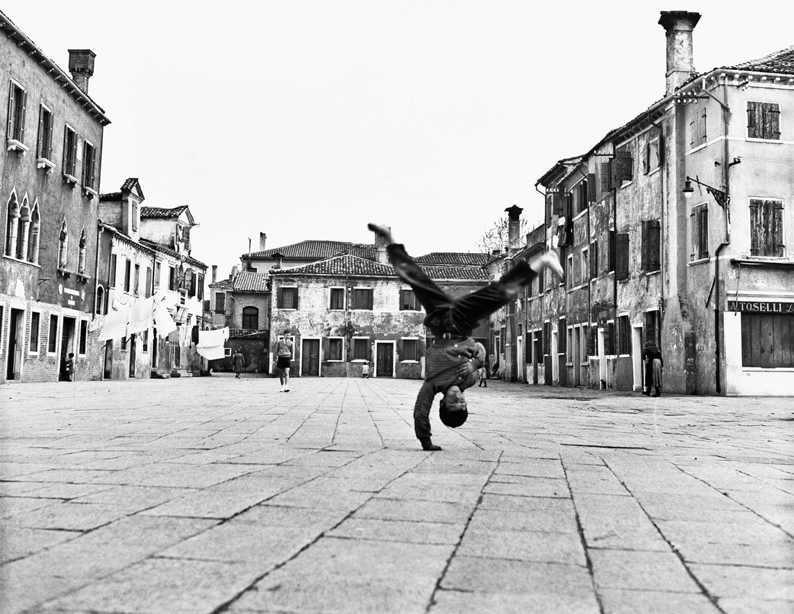 Enfant réalisant une figure de breakdance sur la place de Burano à Venise en 1954, avec des bâtiments anciens en arrière-plan.