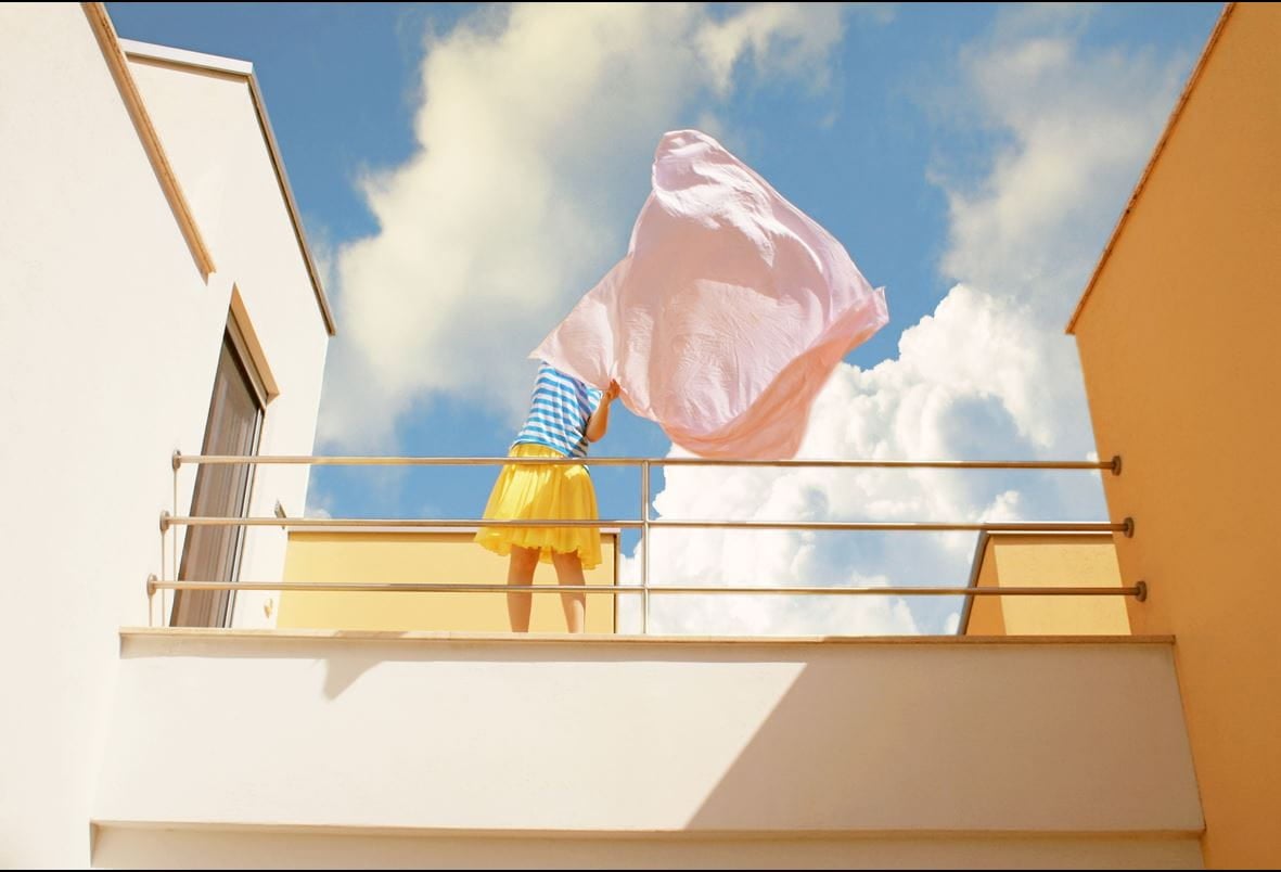 Personne sur un balcon tendant un drap rose sous un ciel bleu avec des nuages.