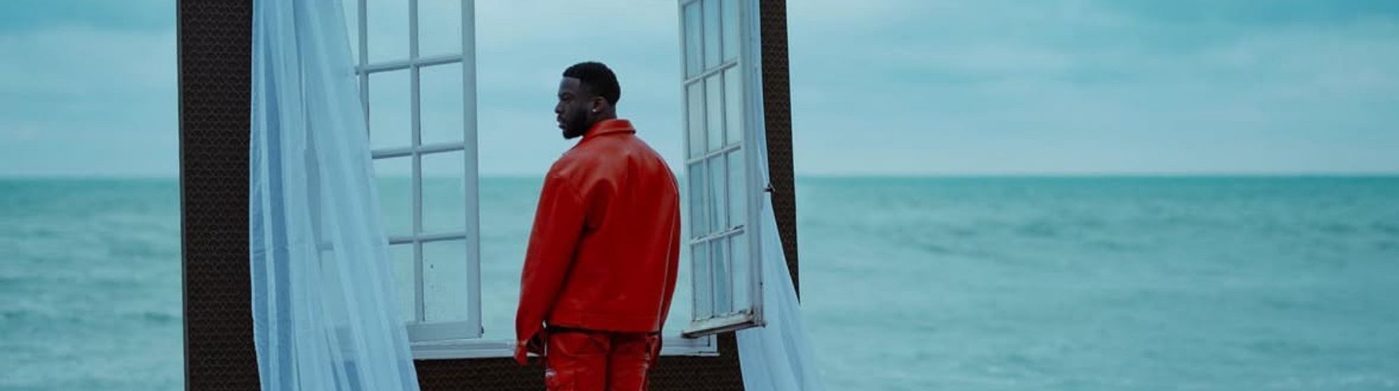 Homme debout sur la plage, vue de dos, vêtu de rouge, entouré d'une structure de fenêtre ouverte avec rideaux blancs