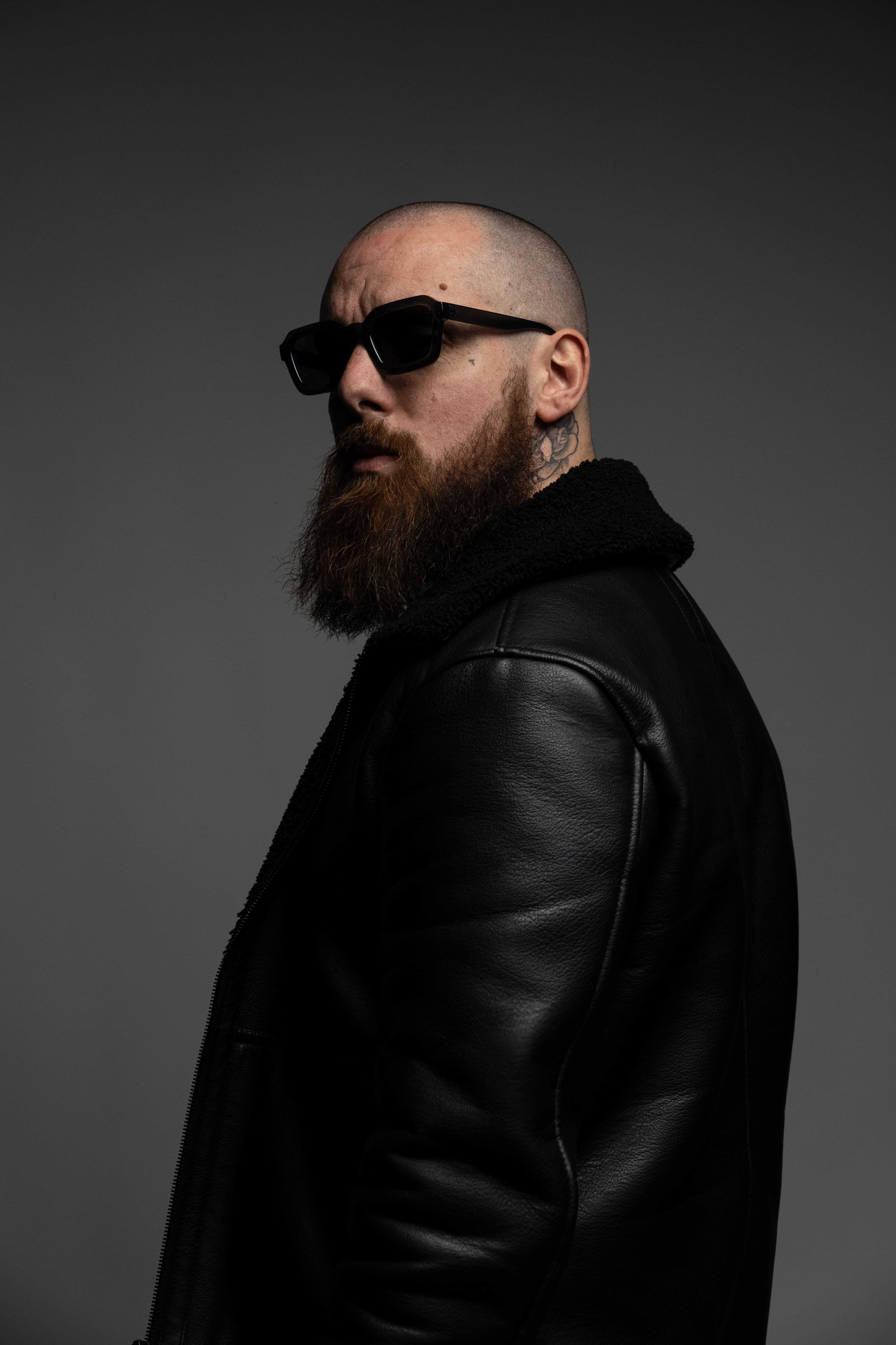 Portrait d'un homme barbu portant des lunettes noires et une veste en cuir, sur fond sombre