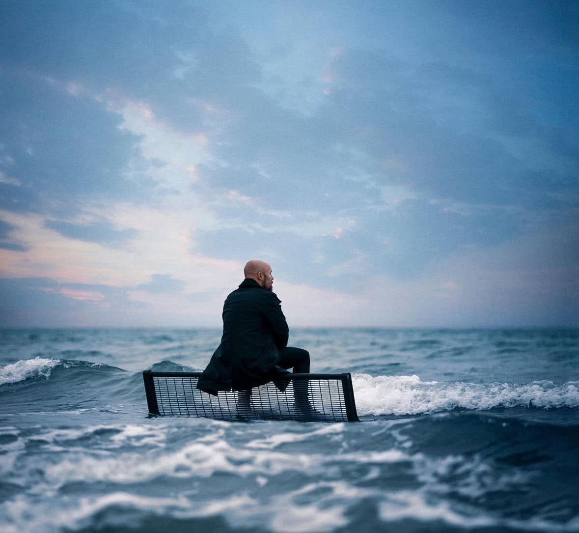 Homme assis sur un banc au milieu de l'océan, habillé en noir, regardant l'horizon sous un ciel crépusculaire