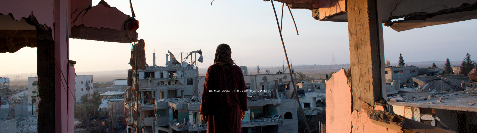Une femme en robe longue regarde les ruines d’une ville détruite à travers l’ouverture d’un bâtiment effondré, au coucher du soleil.