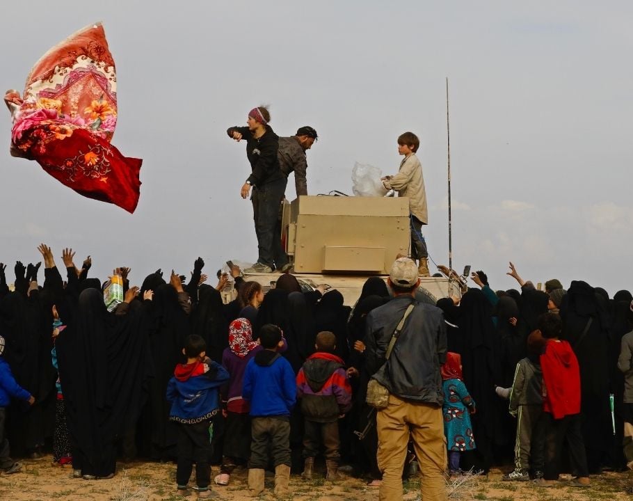 Distribution de couvertures à une foule de réfugiés habillés en noir, entourant un véhicule militaire dans un paysage désertique.