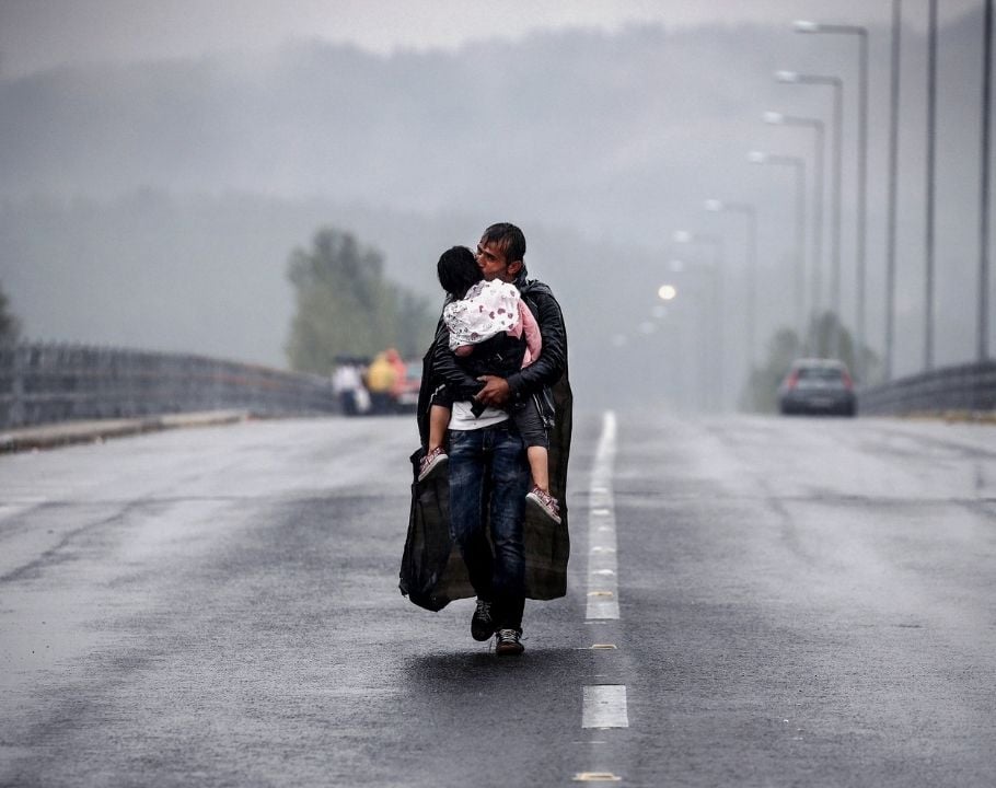 Un père portant son enfant marche seul sur une route mouillée sous la pluie, symbolisant l'exil et la migration.