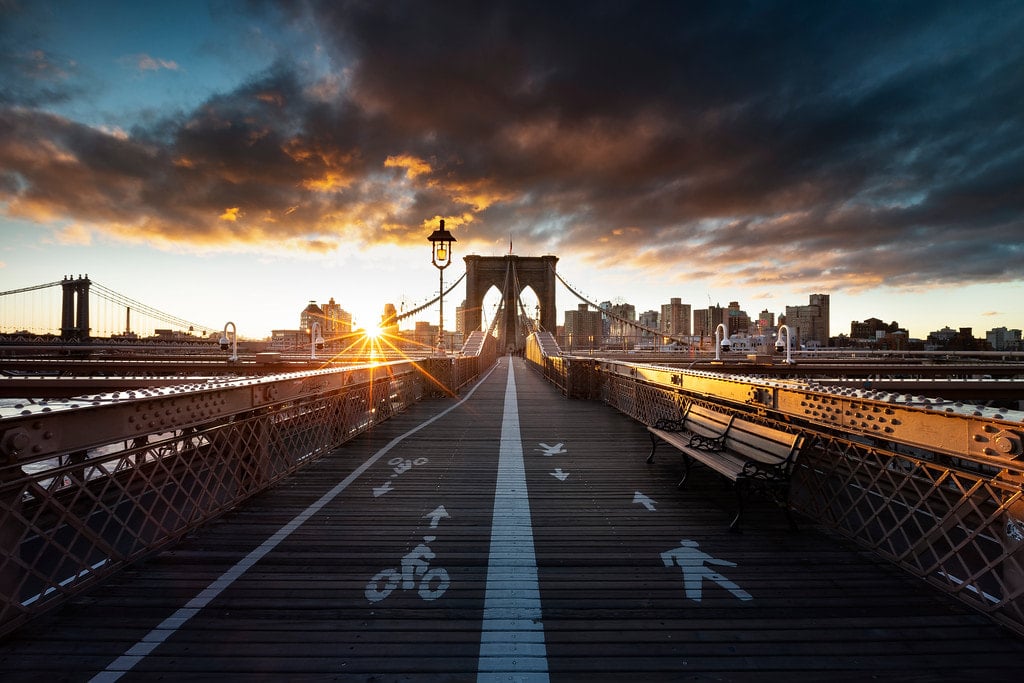 Perspective centrale du pont de Brooklyn au lever du soleil, avec des marquages piétons et cyclistes au sol.