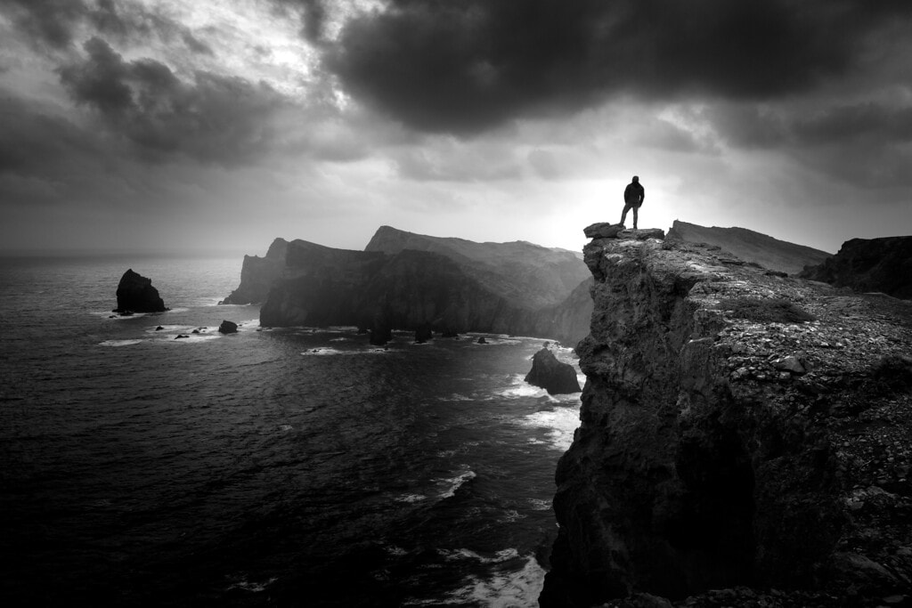 Silhouette d’une personne debout sur une falaise surplombant l’océan, sous un ciel nuageux et contrasté.