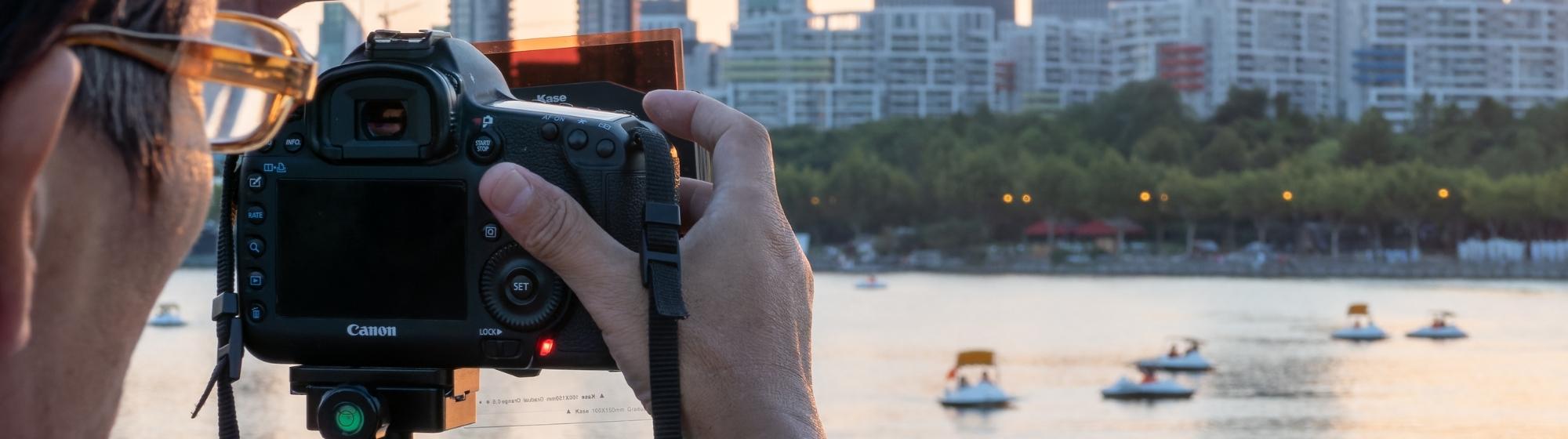 Photographe utilisant un appareil photo équipé d’un filtre optique ND, face à une ville en arrière-plan au coucher du soleil.