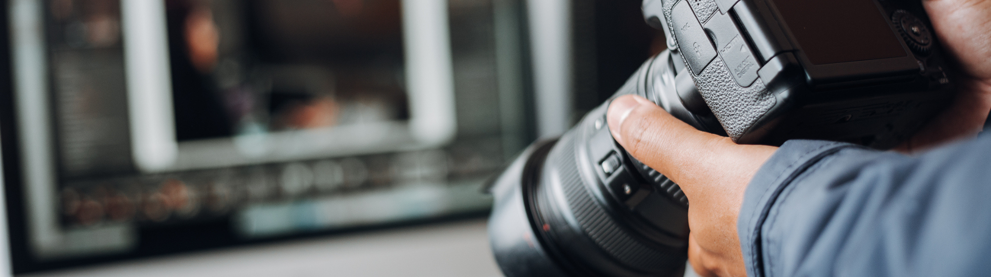 Gros plan sur un photographe tenant un appareil photo devant un écran d’ordinateur affichant un logiciel de retouche.