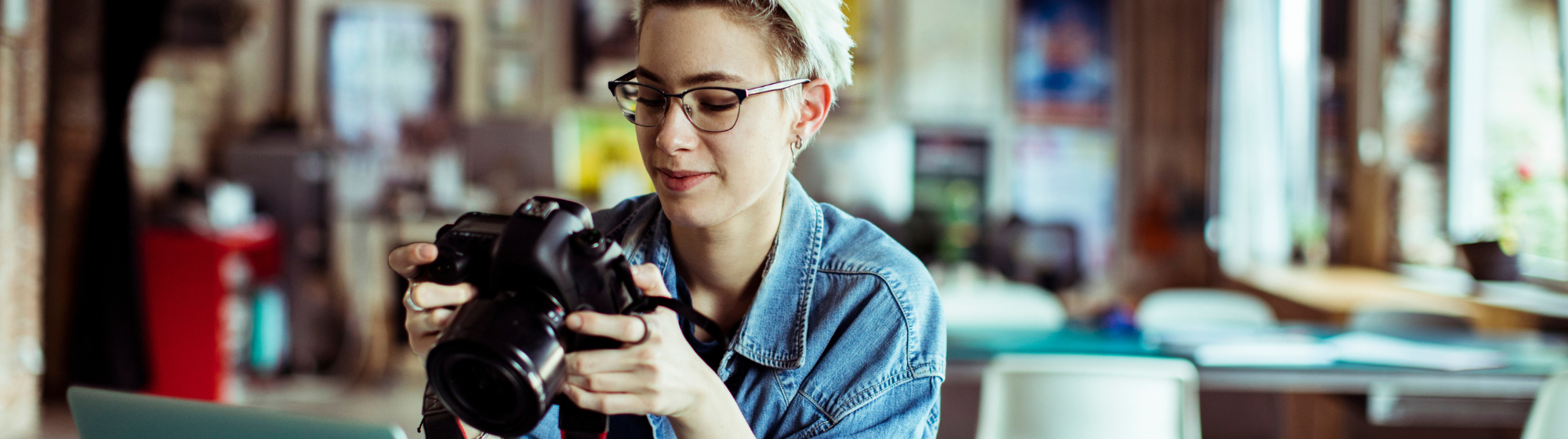 Jeune photographe en chemise en jean consultant son appareil photo devant un ordinateur portable, dans un espace de travail créatif.
