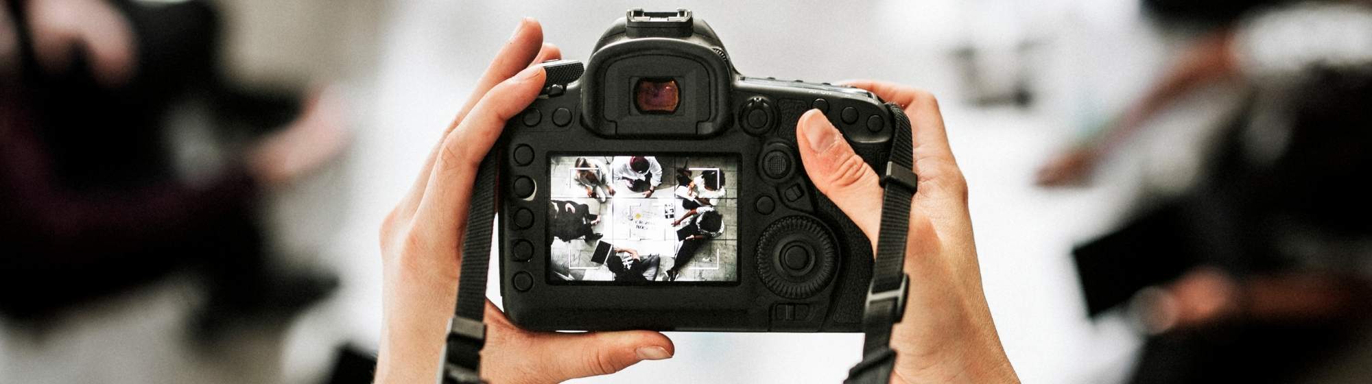 Une personne tenant un appareil photo reflex numérique, prenant une photo d'un groupe de personnes travaillant autour d'une table, visible à travers l'écran de l'appareil photo.