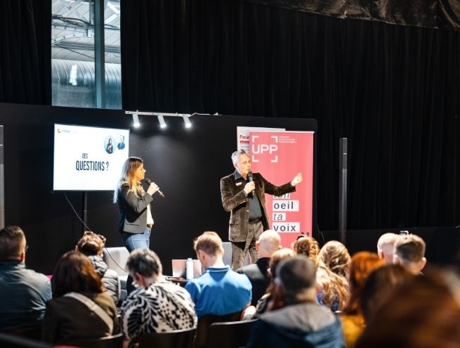 Une scène sur scène avec deux intervenants utilisant des microphones, répondant à des questions devant un public dans un salon ou un événement professionnel.