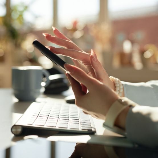 Mains féminines utilisant un smartphone devant un clavier d’ordinateur sur un bureau lumineux.