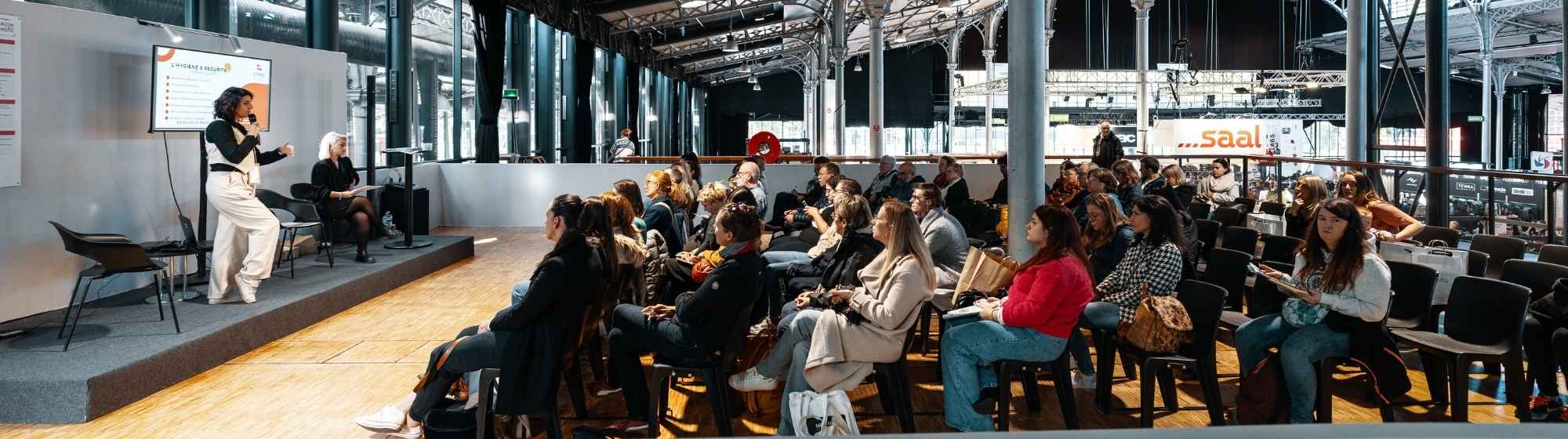 Vue panoramique d’une conférence en salon avec une intervenante sur scène et un large public attentif.