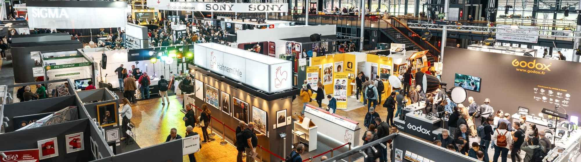 Vue d'ensemble d'un hall d'exposition animé avec des stands et des présentations, et de nombreuses personnes qui se promènent et interagissent dans un grand espace ouvert.