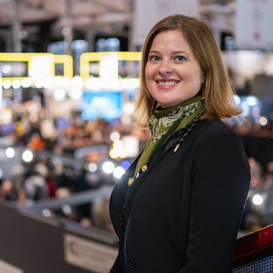 Portrait de Marianne souriante, photographiée lors d'un salon professionnel, avec les stands en arrière-plan.